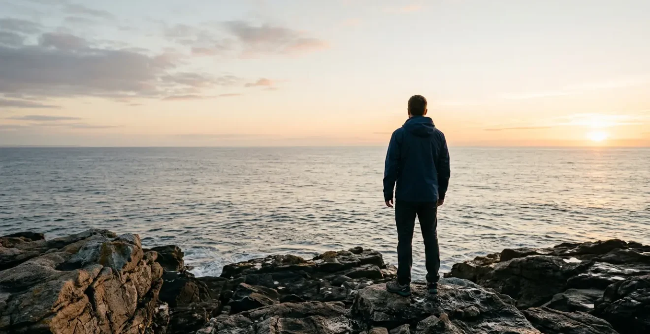 A person standing at the edge of a rocky coastal cliff at dawn, looking toward a bright horizon over calm ocean waters, symbolizing hope and new beginnings after professional setback