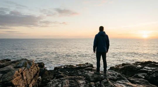 A person standing at the edge of a rocky coastal cliff at dawn, looking toward a bright horizon over calm ocean waters, symbolizing hope and new beginnings after professional setback